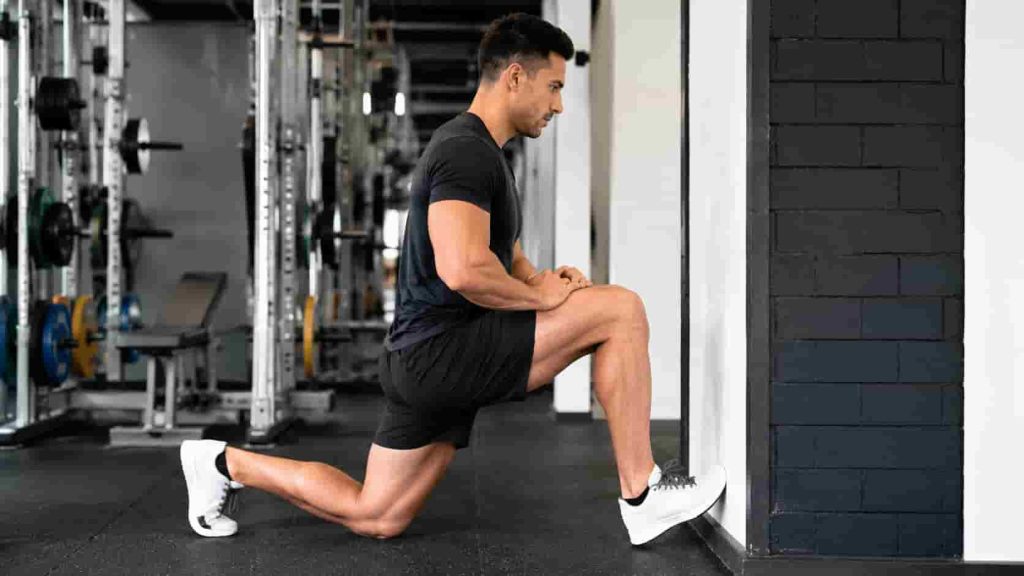 A man in a gym doing a knee-to-wall ankle mobility stretch to improve dorsiflexion and range of motion.