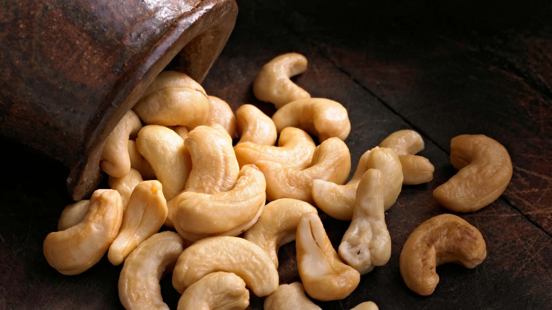 Whole raw cashews spilling out of an overturned rustic brown ceramic jar onto a dark textured wooden surface.