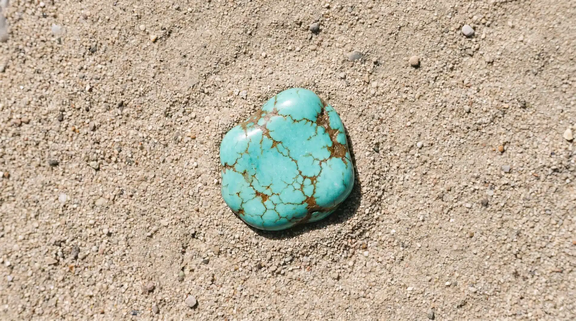 A polished blue-green turquoise stone with brown veins resting on a sandy, textured surface.