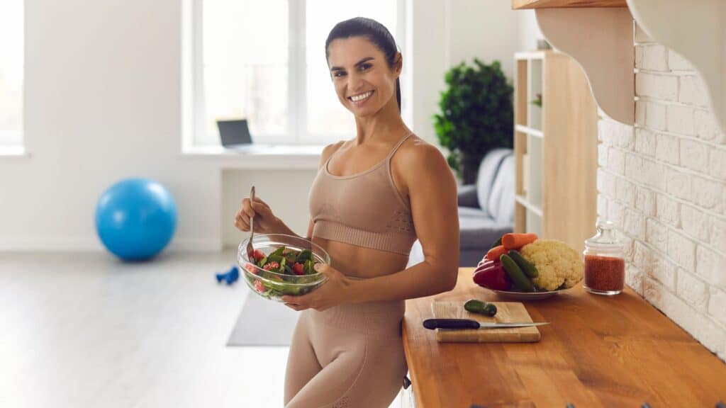 A smiling woman in athletic wear holding a bowl of fresh salad in a kitchen.