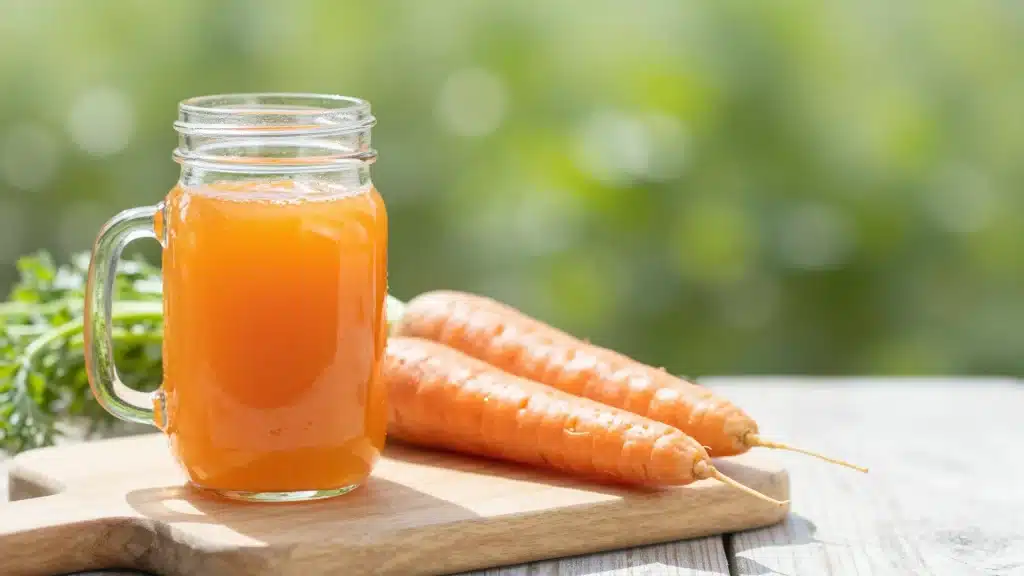 Alt text: Fresh carrot juice in a glass mason jar next to whole carrots on a cutting board.