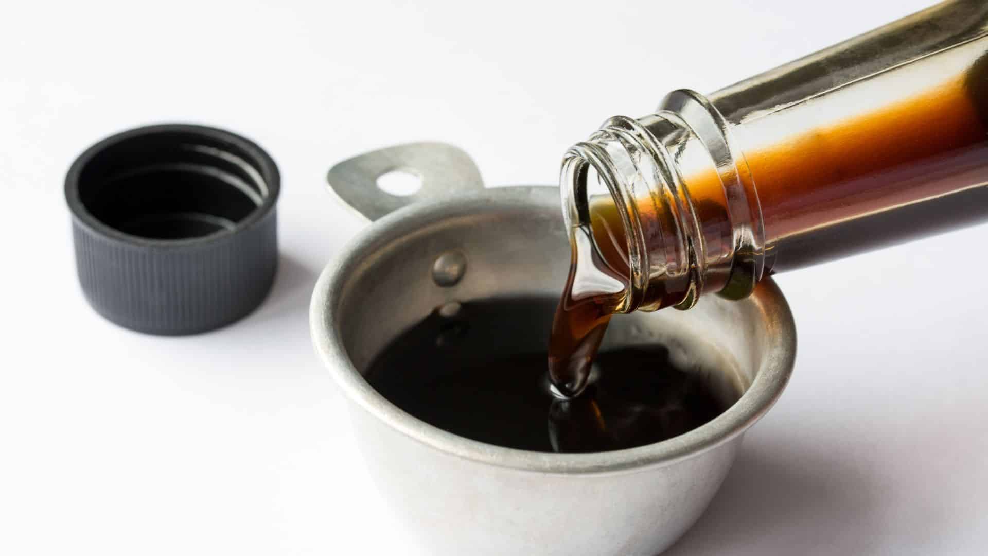 Dark coconut aminos pouring from a clear glass bottle into a small stainless steel measuring cup on a white surface.
