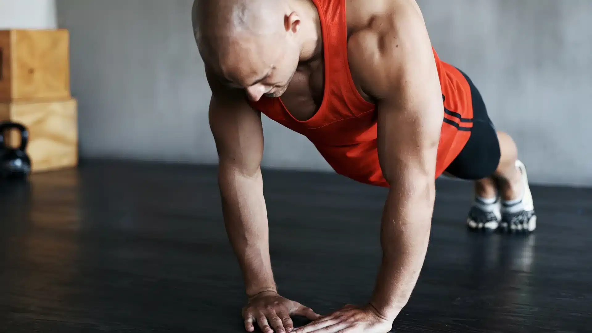 A side-view of a man in a red tank top performing a diamond push-up on a dark gymnasium floor.
