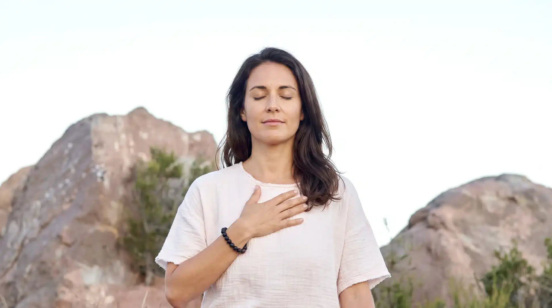 A woman in a white shirt sitting with her eyes closed in meditation, holding one hand over her chest against a rocky backdrop.