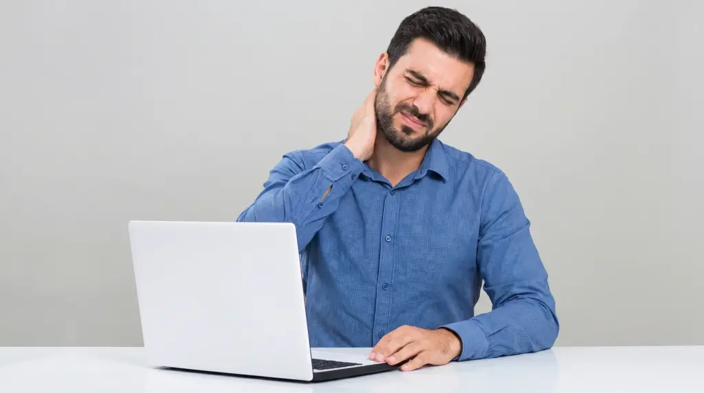 A man in a blue shirt sitting in front of a laptop, grimacing and holding his neck in pain.