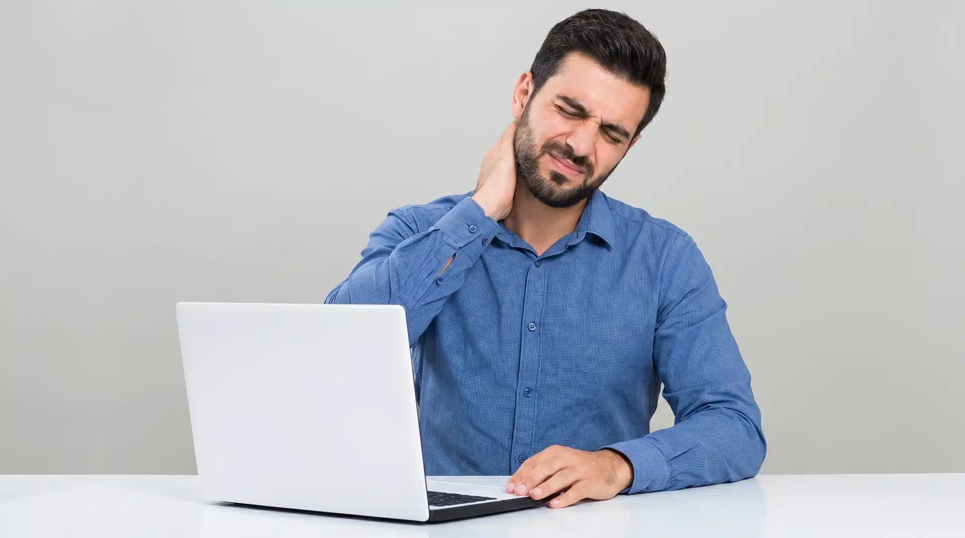 A man in a blue shirt sitting in front of a laptop, grimacing and holding his neck in pain.