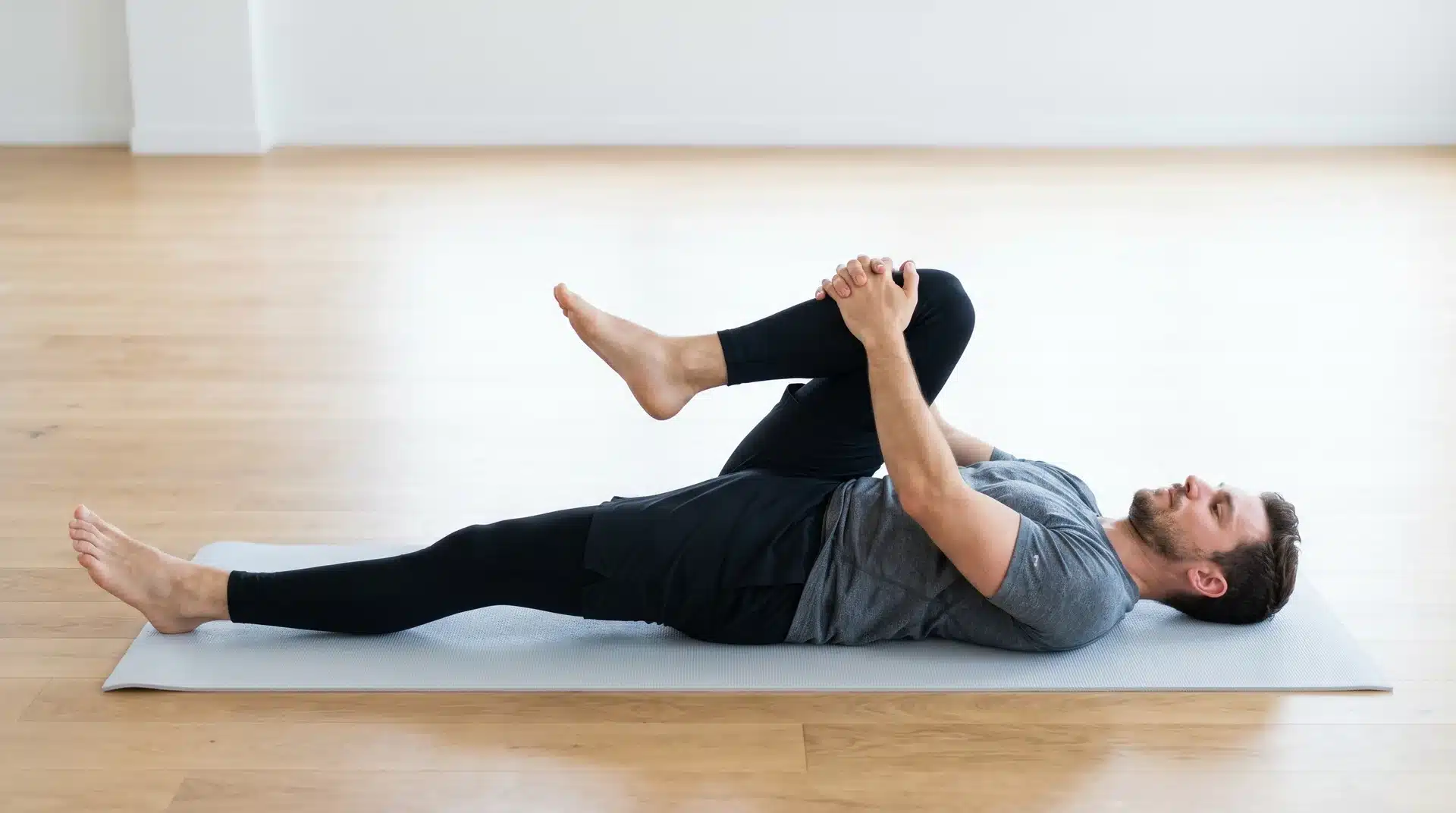 A side-view of a man in athletic wear performing a knee-to-chest stretch on a white mat, pulling one knee to his torso.
