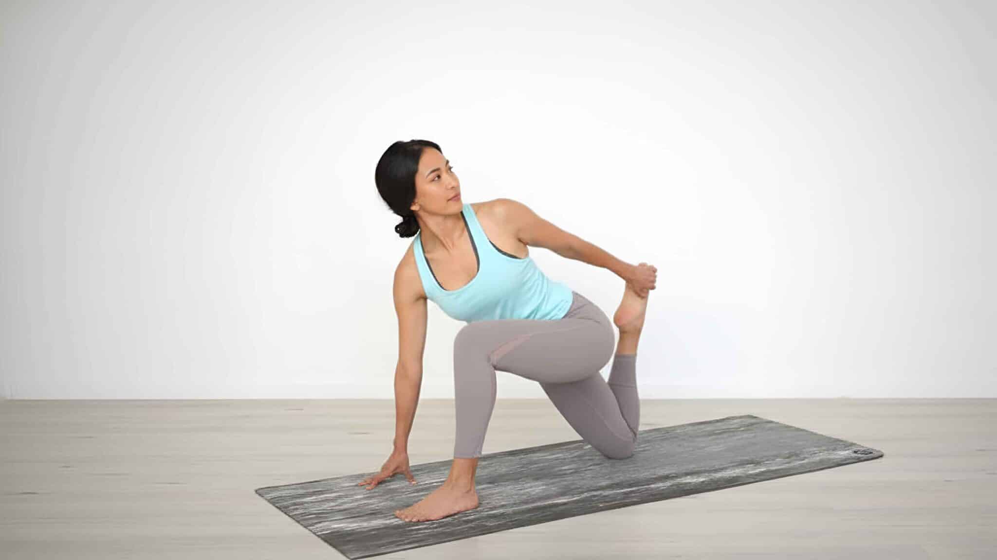 A woman on a yoga mat in a kneeling low lunge position, reaching back with her left hand to pull her left foot toward her glutes for a kneeling quad stretch.