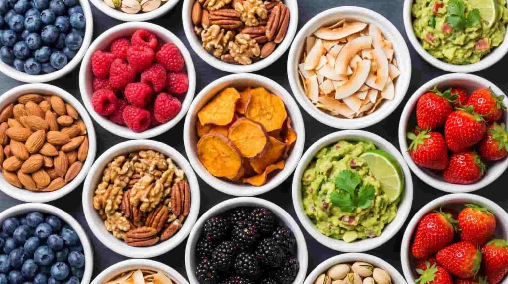 Assorted bowls of paleo snacks including fresh berries, almonds, walnuts, pumpkin chips, and guacamole dip.