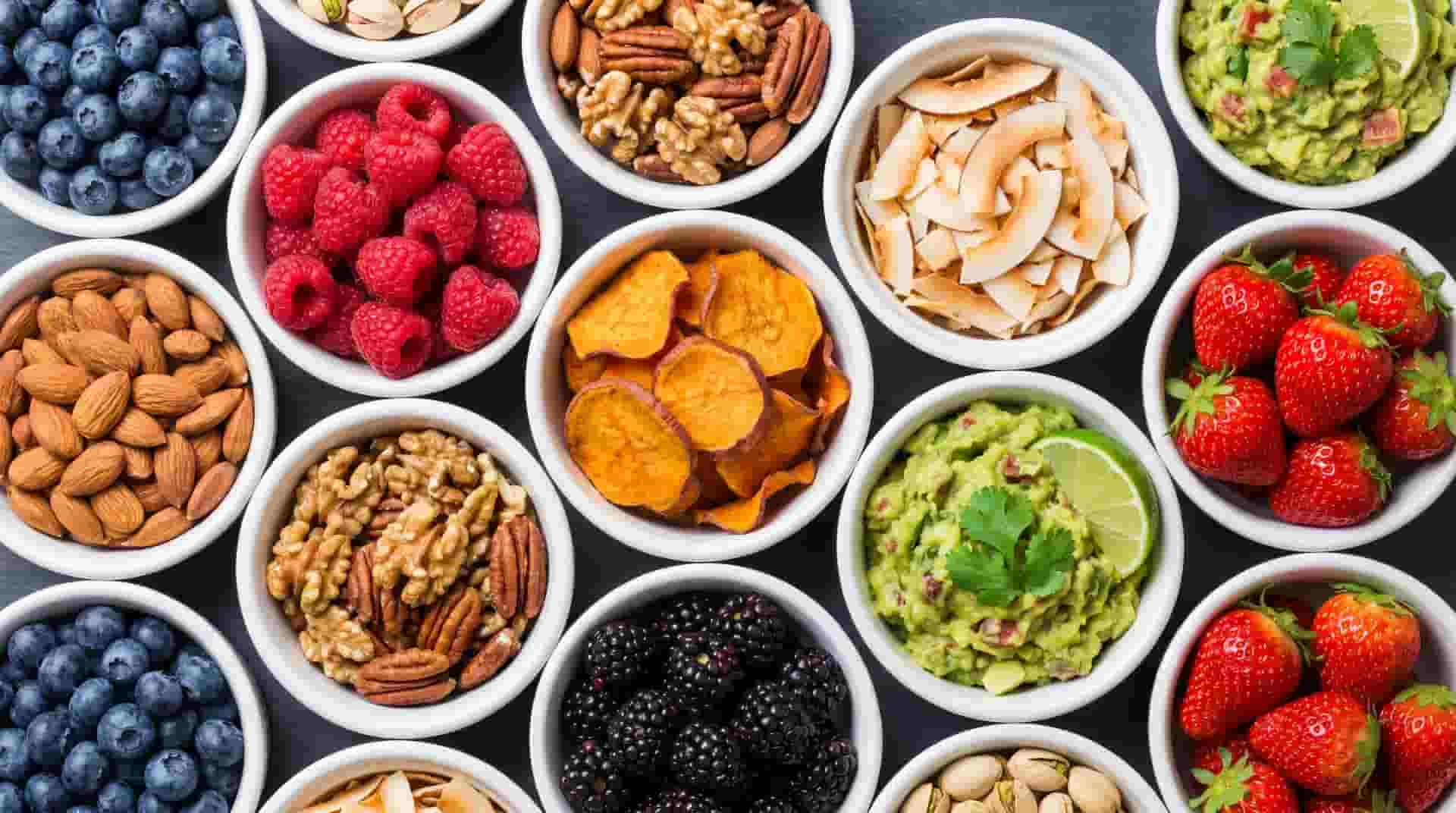 Assorted bowls of paleo snacks including fresh berries, almonds, walnuts, pumpkin chips, and guacamole dip.