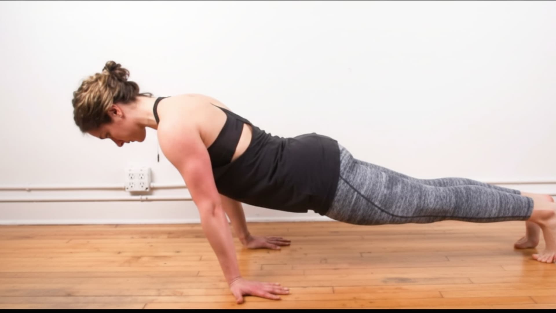 A side-view of a woman in black athletic wear performing a pseudo planche push-up hold on a light-colored wooden floor.