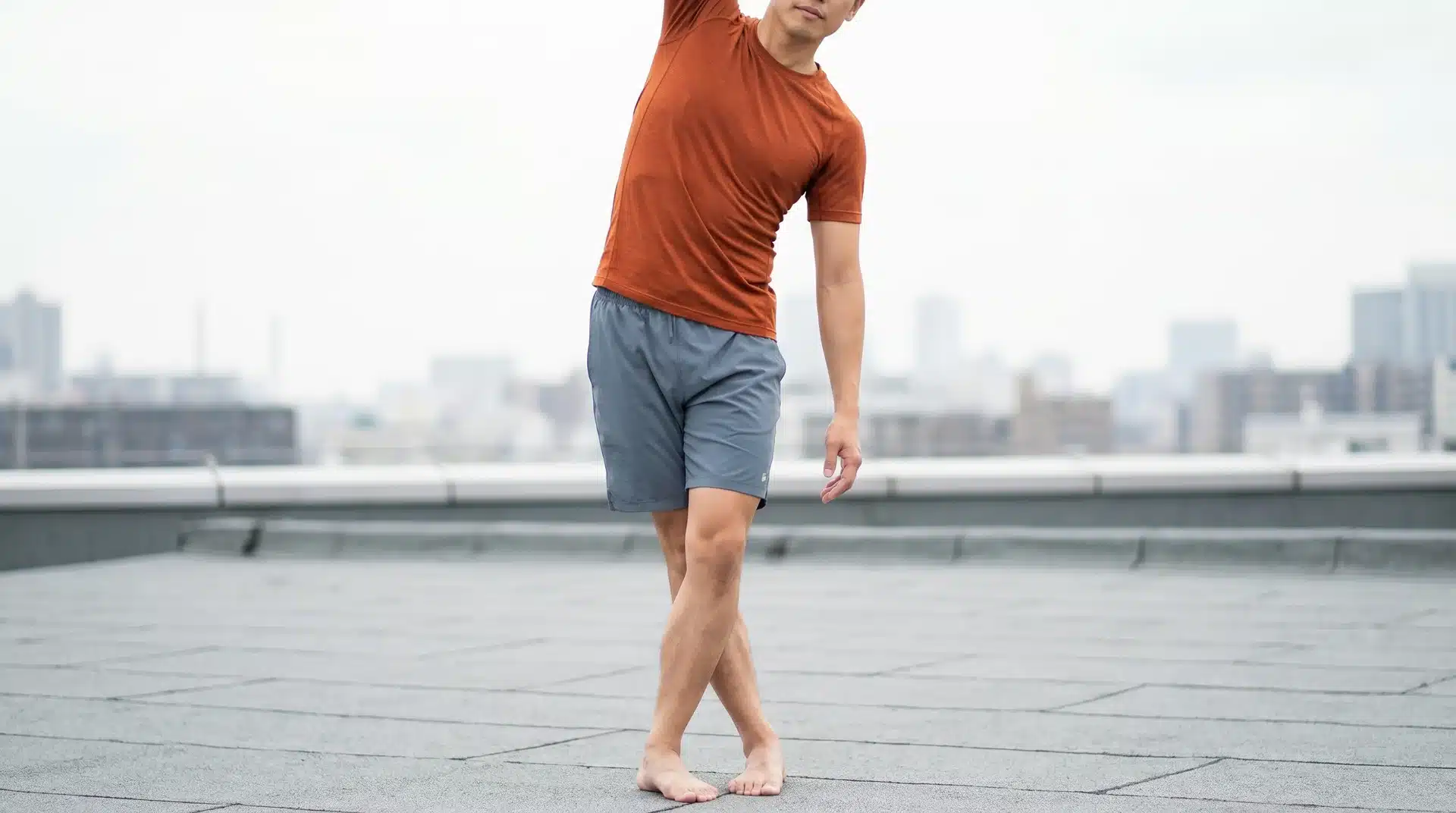 A man standing outdoors, crossing his legs and leaning sideways with one arm overhead to stretch his outer thigh.