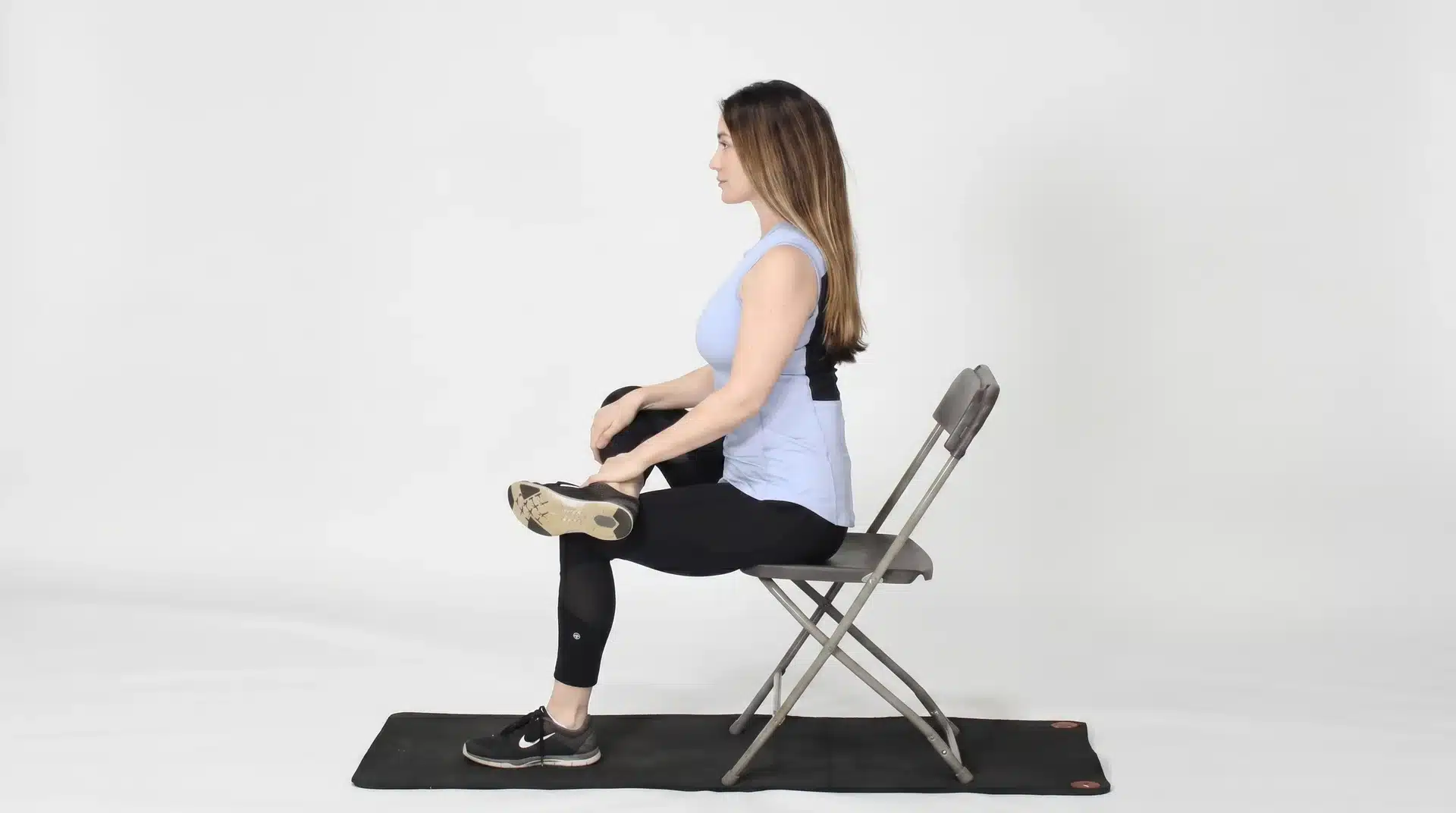A side-view of a woman in athletic wear sitting upright in a metal folding chair, performing a seated cross-leg hip stretch.