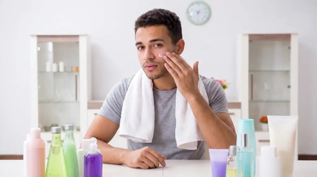 A young man applying moisturizer to his face in front of a bathroom mirror, with skincare products on the counter.