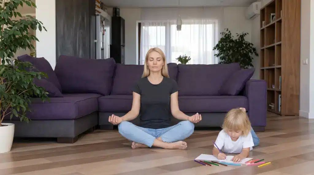 A mother practicing meditation in a lotus pose on the living room floor while her young toddler sits beside her drawing with crayons.