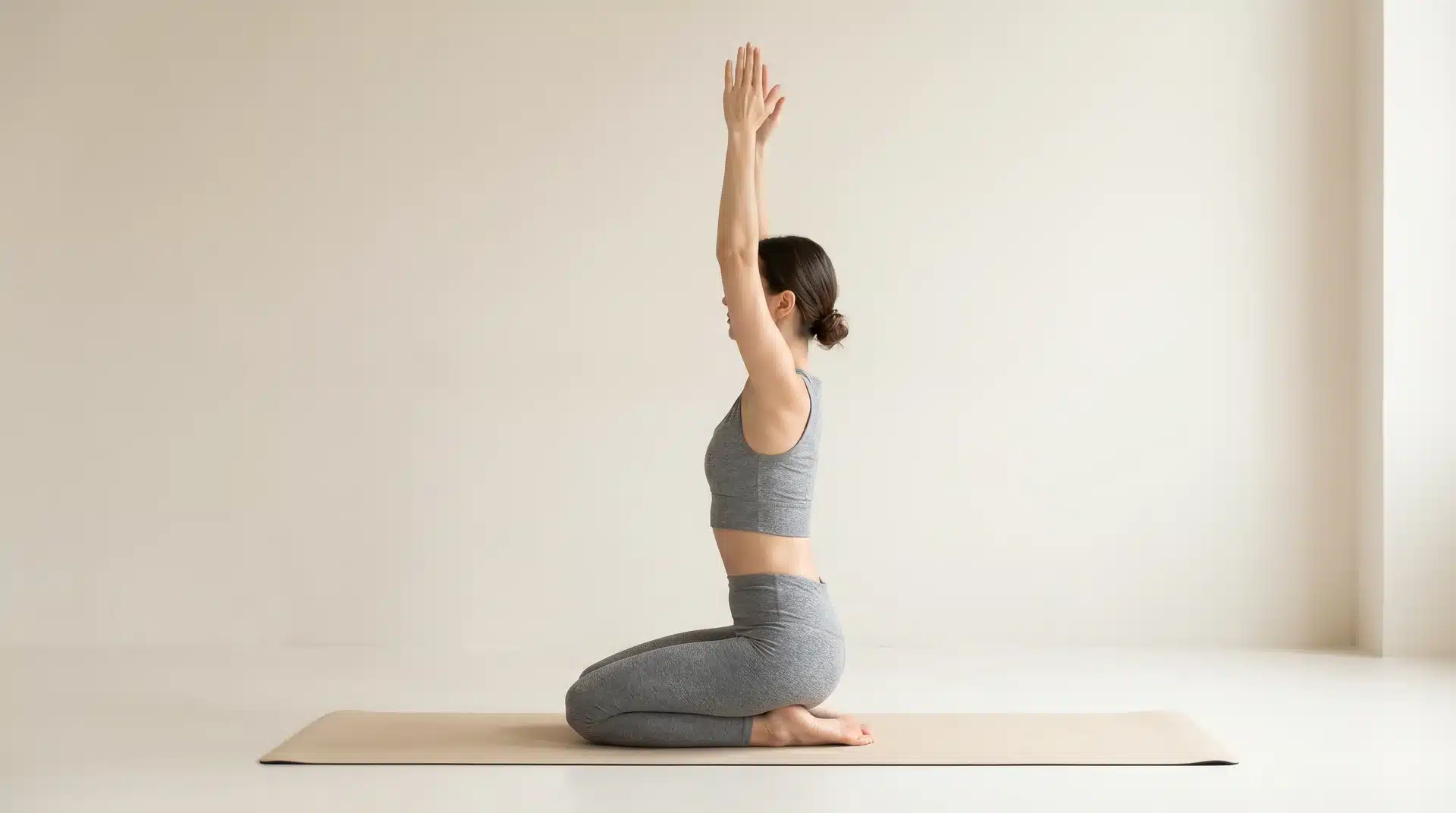 Side view of a woman kneeling in Seiza pose on a mat with both arms extended straight up overhead.