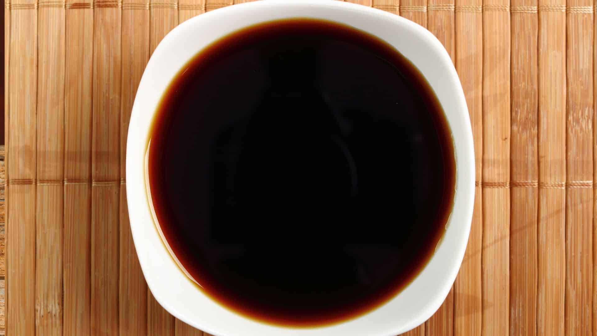 A top-down view of dark soy sauce in a white ceramic bowl, sitting on a natural light-brown bamboo placemat.