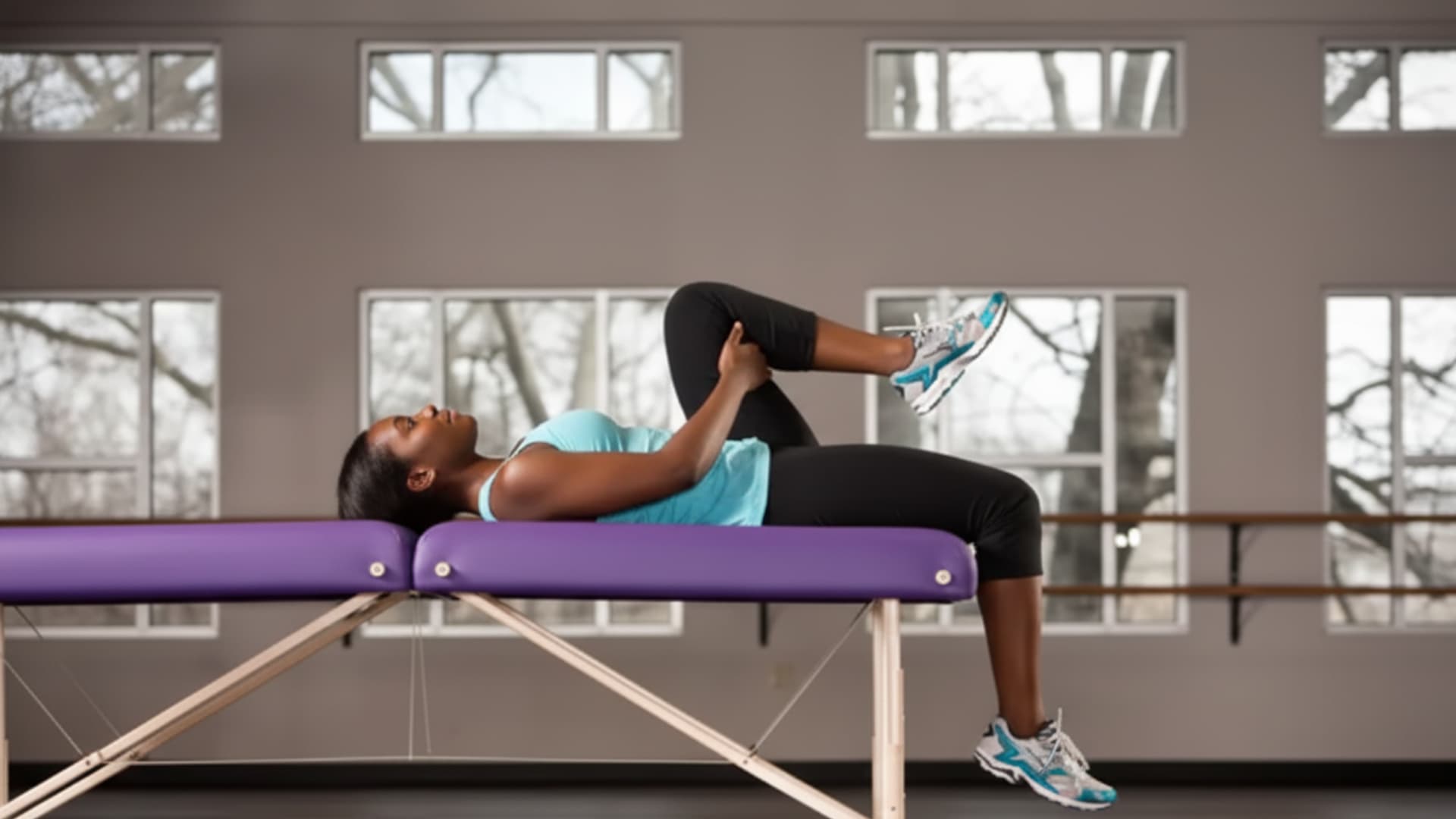 A woman lying on her back on a massage table, pulling one knee to her chest while letting her other leg hang off the edge.