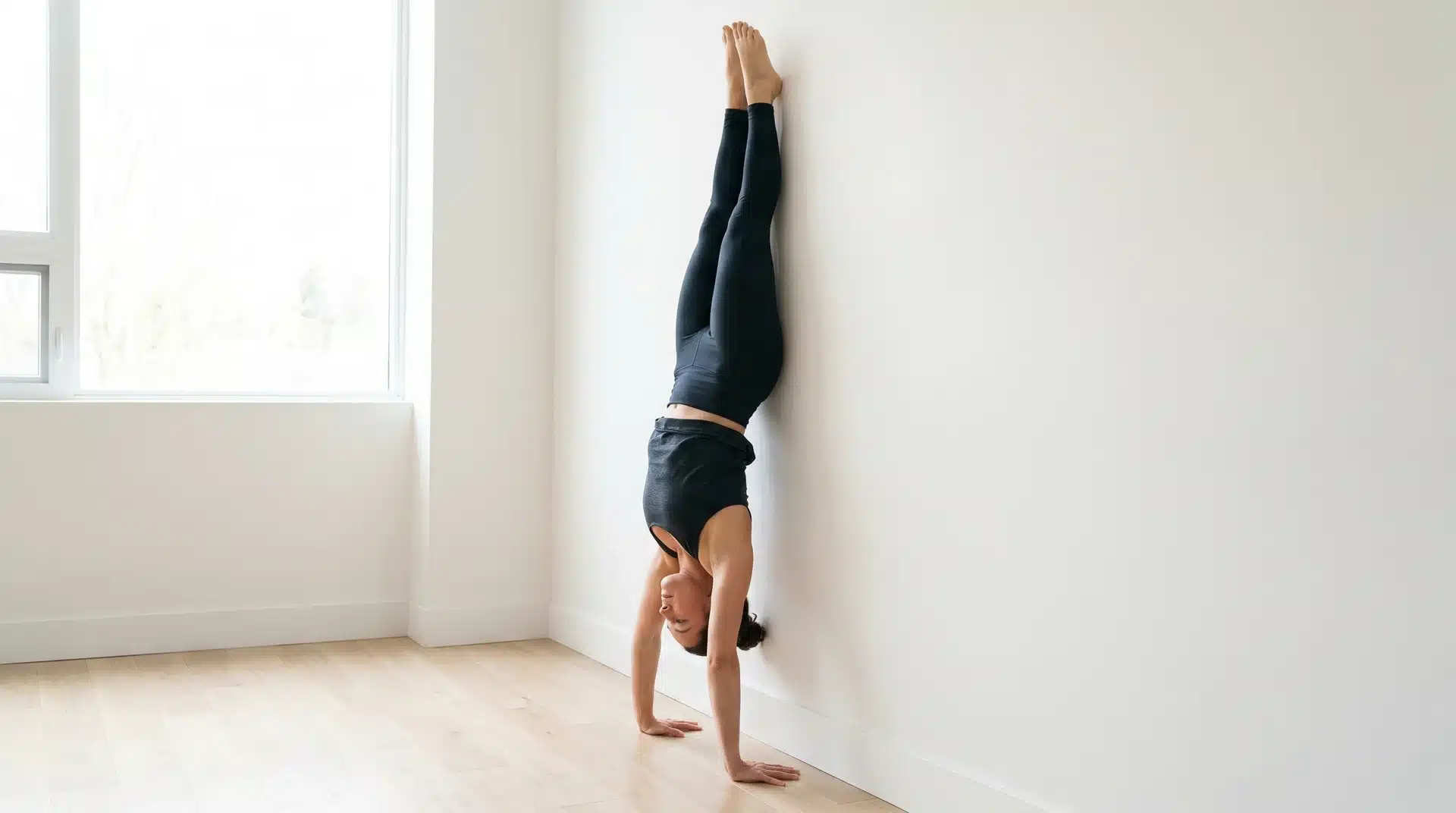 A side-view of a person in athletic wear performing a vertical wall handstand hold in a bright studio.