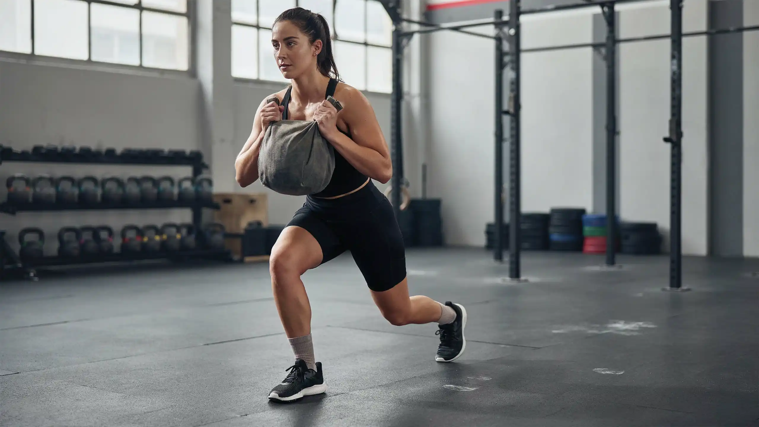 Female athlete completing sandbag lunges during HYROX training, focusing on lower body strength, balance, and race-specific conditioning.