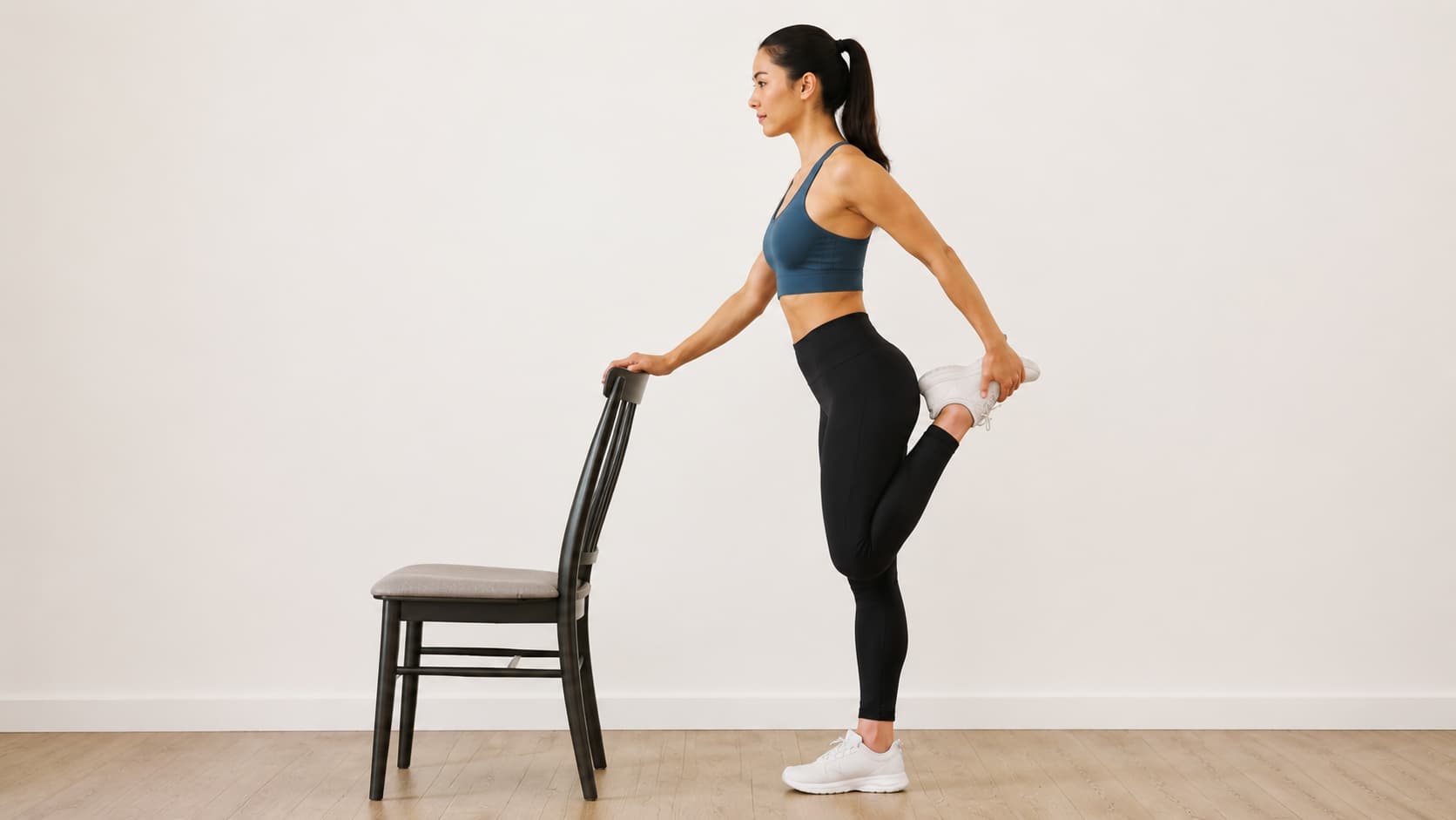 Woman in profile view holding the back of a wooden chair for balance while performing a standing quad stretch.