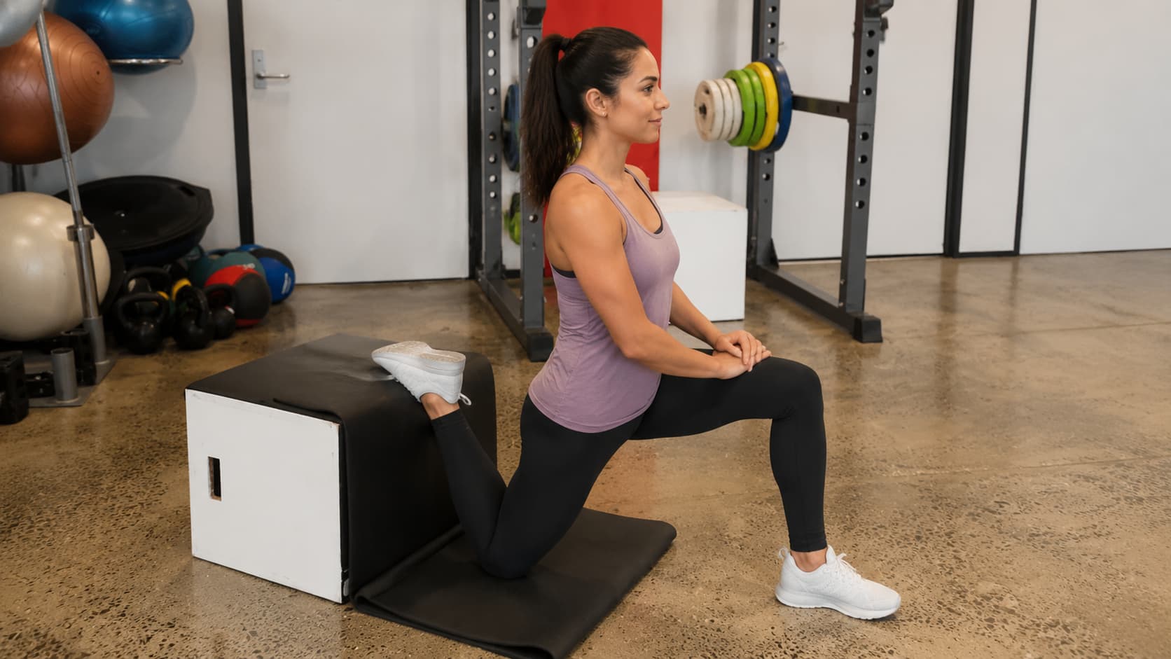 Woman on a mat performing a kneeling quad stretch with her back foot elevated on a black fitness bench.