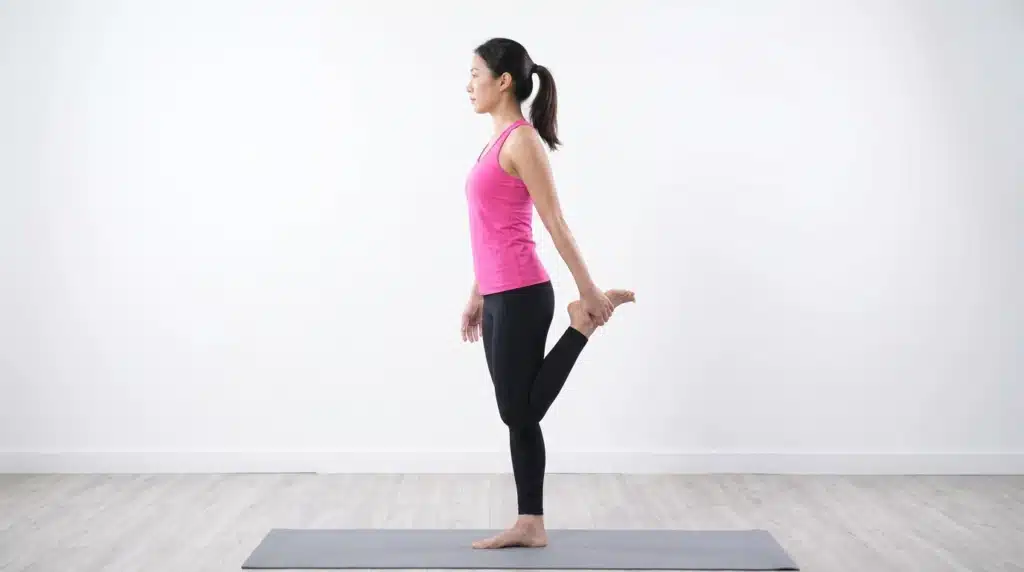 A woman in a pink tank top and black leggings stands on a yoga mat, performing a standing quad stretch by holding her left ankle behind her.