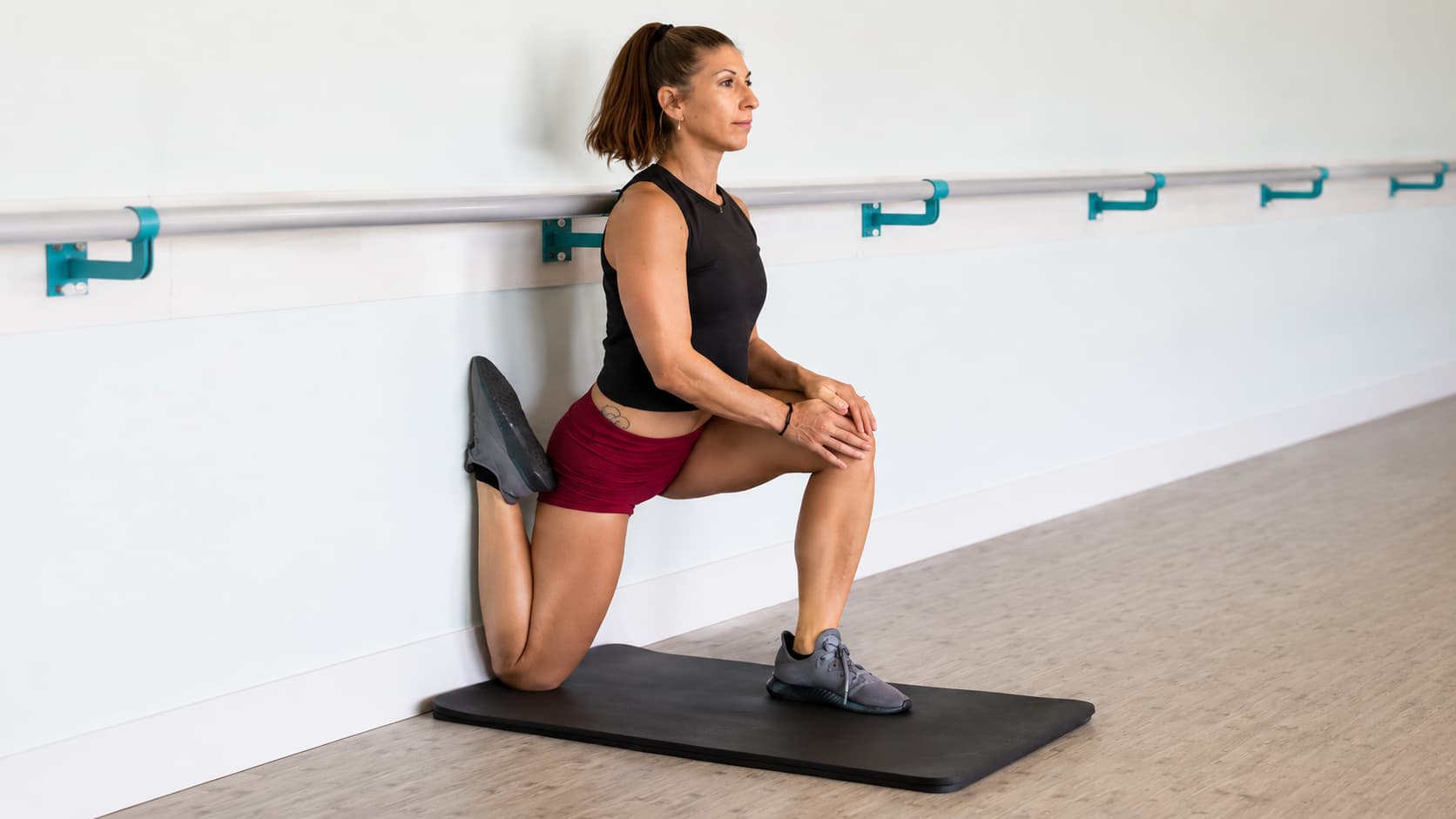 Woman on a mat performing a wall-supported kneeling quad stretch with her back shin pressed flat against a wall.