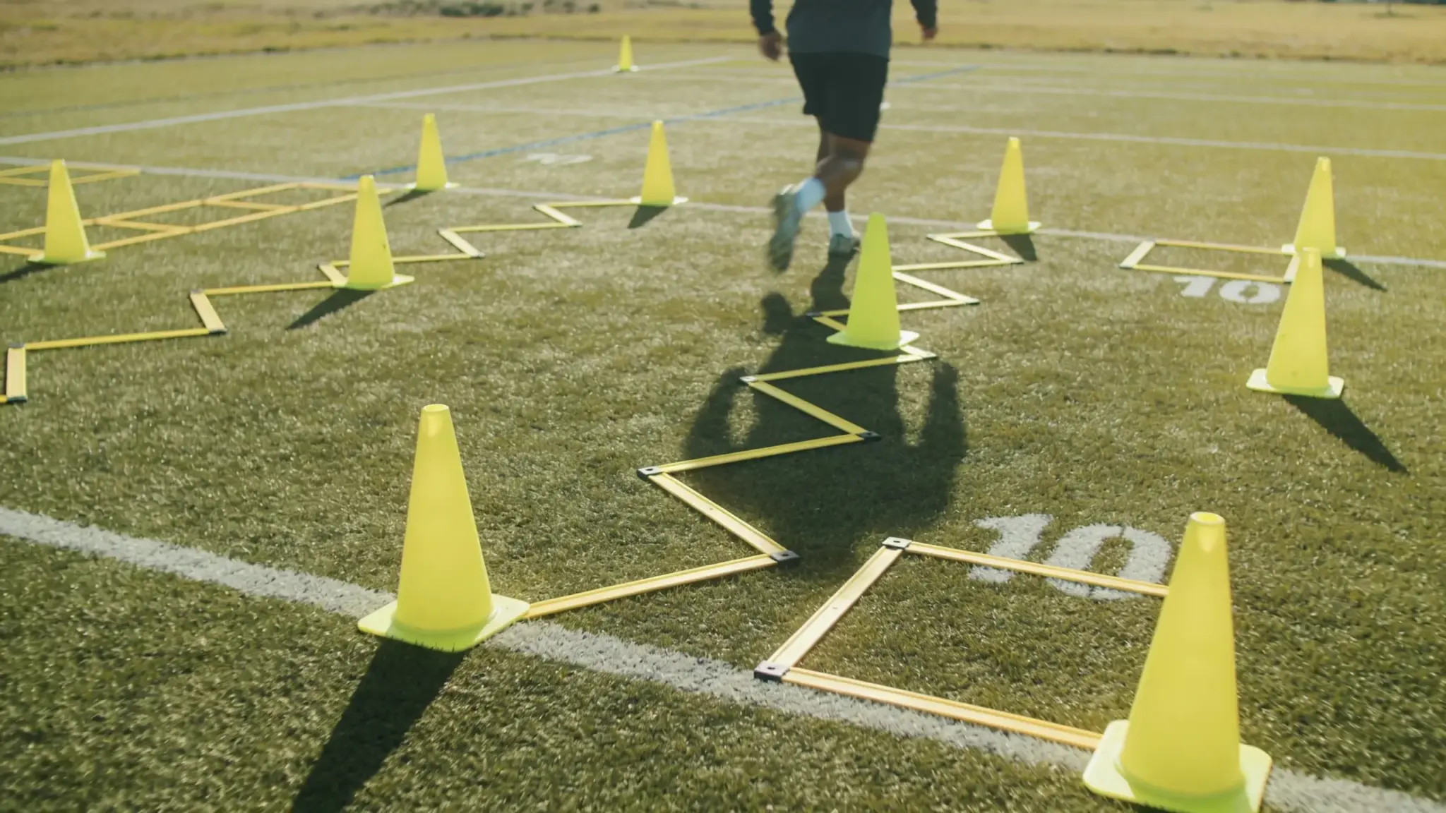 Athlete performing agility drill on sports field using yellow cones and ladder markers during outdoor training in sunlight