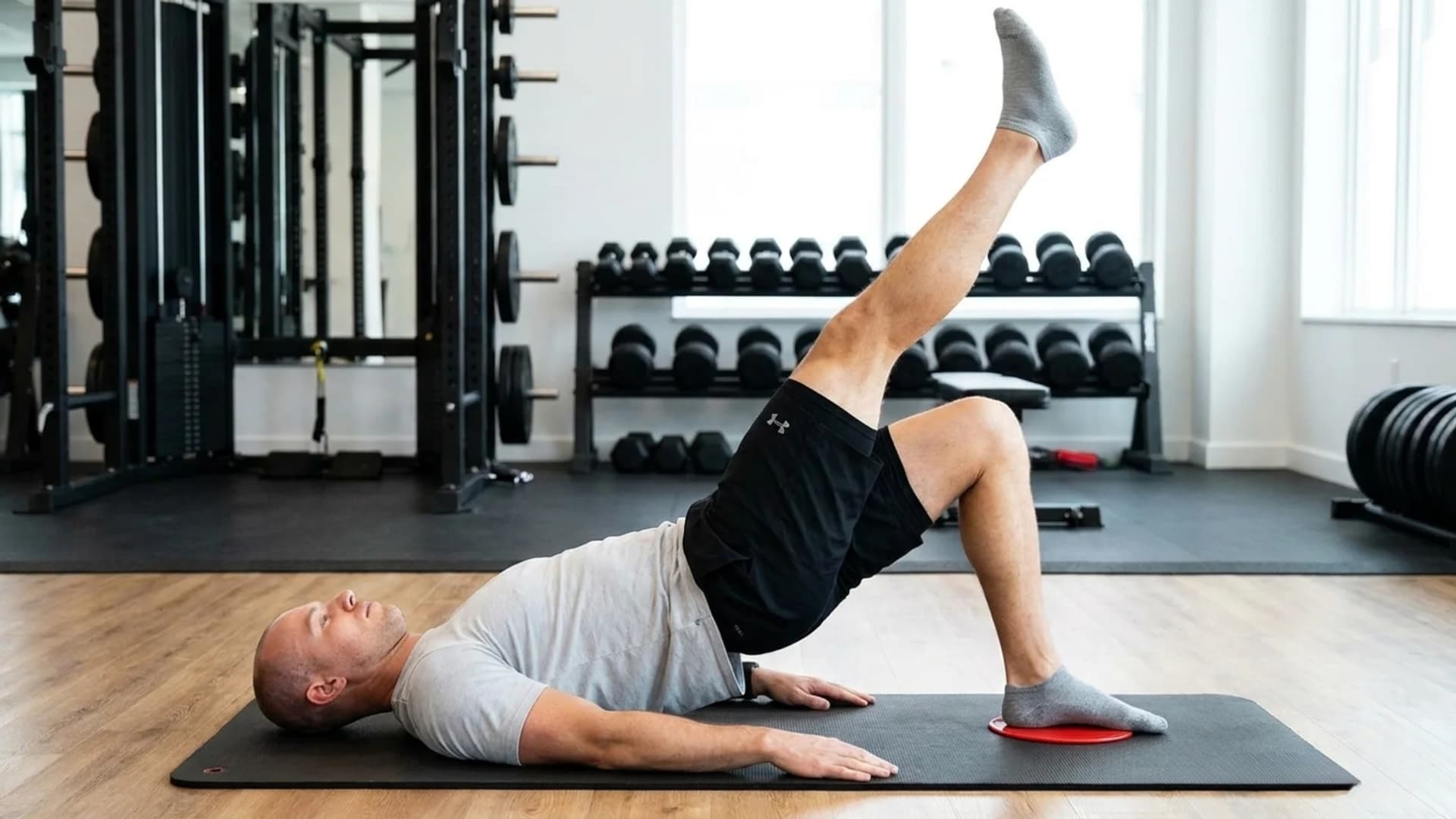 Man performing a single-leg eccentric hamstring bridge exercise on an exercise mat using a sliding disc.